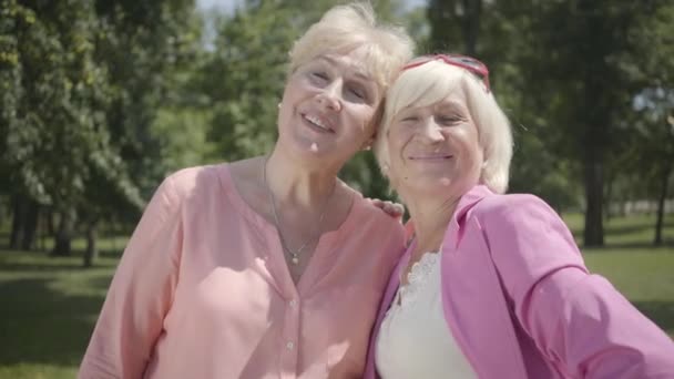 Retrato de dos ancianas positivas mirando a la cámara sonriendo de pie en el parque de verano. Novias de ocio al aire libre. Señoras maduras descansando juntas en el jardín — Vídeos de Stock