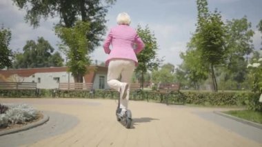 Parkta Binicilik sırasında bir scooter kullanarak pembe ceket neşeli sevimli kıdemli kadın. Modern kadın, yeni nesil. Sağlıklı neşeli kıdemli emekli bayan. Ekolojik ulaşım konsepti