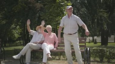 İki kıdemli erkek ve bir kadın el sallıyor, arkadaşı kadın parkta scooter sürerken. Olgun insanlar açık havada istirahat, aktif yaşam tarzı. Neşeli kıdemli emekli insanlar.
