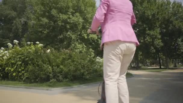 Mujer mayor positiva alegre en chaqueta rosa usando una scooter mientras monta en el parque. Mujer moderna, una nueva generación. Saludable señora jubilada alegre. Concepto de transporte ecológico — Vídeo de stock