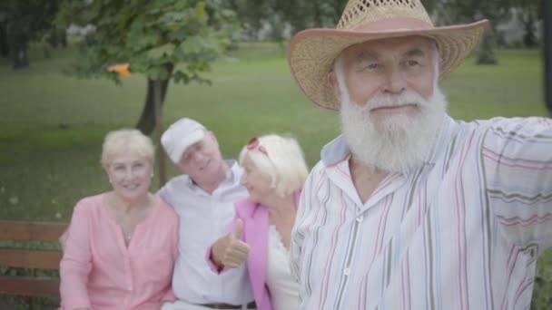 Vuxen man i en hatt och grått skägg gör en selfie mot bakgrunden av sina vänner sitter på en bänk i parken. Två bedårande Joy par vilar utomhus tillsammans. Glad Senior pensionerad — Stockvideo