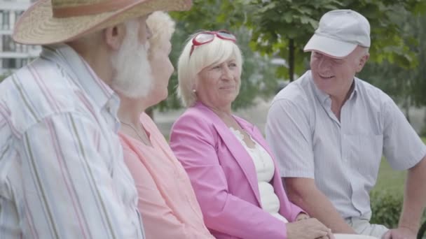 Två söta mogna par pratar och leende sitter på bänken i sommar parken. Dubbel datum för seniora par. Vänligt sällskap som vilar utomhus. Gamla män och kvinnor träffades tillsammans. Pensionerade personer. — Stockvideo