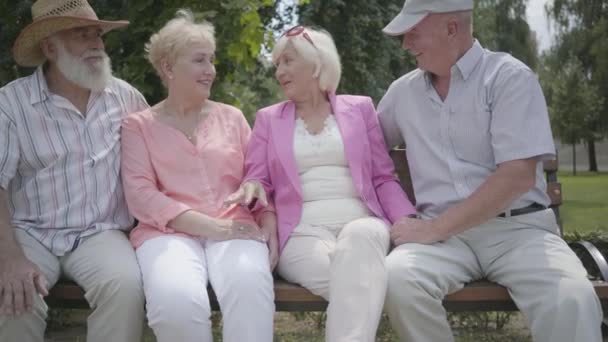 Två söta mogna par pratar och leende sitter på bänken i sommar parken. Dubbel datum för seniora par. Vänligt sällskap som vilar utomhus. Gamla män och kvinnor träffades tillsammans. Pensionerade personer. — Stockvideo