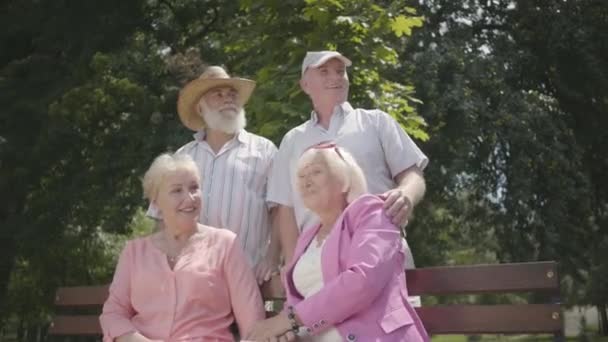 Två bedårande mogna par pratar och ler vid bänken i sommar parken. Dubbel datum för seniora par. Vänligt sällskap som vilar utomhus. Gamla män och kvinnor träffades tillsammans. Pensionerade personer. — Stockvideo