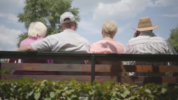 Två bedårande mogna par sitter på bänken och pratar tillsammans. Mogna människor som vilar utomhus. Glada pensionärer. Back View. Slow motion. — Stockvideo