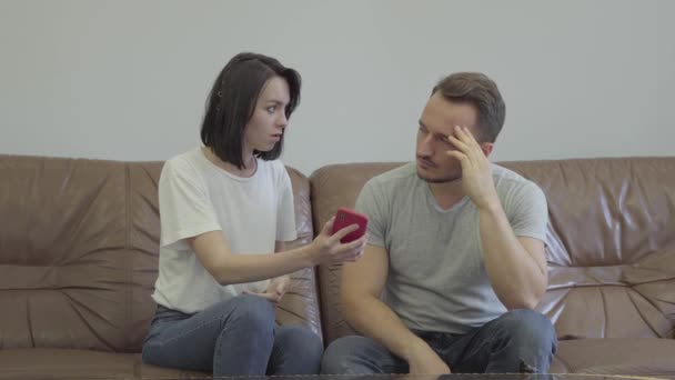 Portrait of angry woman shouting at her sad husband showing cellphone with messages from his paramour at home. Problems in the relationship between man and woman. Betrayal, mistrust, breakup concept — Stock Video