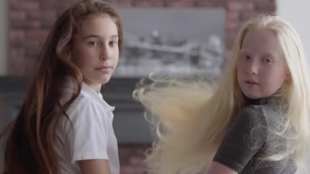 Två söta tjejer med långt vackert hår sitter nära varandra i vardagsrummet. Begreppet vänskap. Bekymmerslös barndom. Bedårande flickor vända och titta på kameran. Real People-serien. — Stockvideo