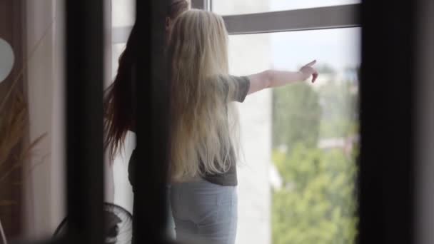 Due bambine con i capelli biondi e scuri che guardano fuori dalla finestra nel soggiorno. Ragazza bruna e ragazza albina con i capelli lunghi che giocano a casa. Infanzia spensierata. Rallentatore . — Video Stock