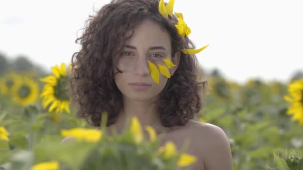 Portret van schattig krullend speels meisje kijken naar de camera glimlachend staande in de zonnebloem veld. Heldere gele kleur. Het meisje bedekt zichzelf met zonnebloemen. Freedom concept. Gelukkige vrouw buitenshuis — Stockvideo