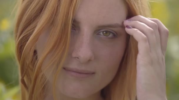 Gros plan sur le visage d'une fille sensuelle confiante aux cheveux roux et aux yeux verts debout sur le champ de tournesol en regardant la caméra. Concept de beauté, lien avec la nature . — Video