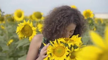 Sevimli kıvırcık oyuncu kadın kamera ayçiçeği alanında ayakta gülümseyerek bakarak portresi. Parlak sarı renk. Elinde ayçiçeği buketi olan kız. Mutlu kadın açık havada