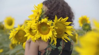 Oldukça kıvırcık oyuncu kız portresi ayçiçeği alanında ayakta gülümseyerek kameraya bakarak. Parlak sarı renk. Elinde ayçiçeği buketi olan kız. Mutlu kadın açık havada