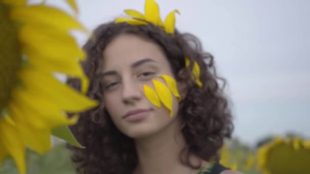 Porträtt av vackra Curly lekfull flicka tittar på kameran stående på solros fältet. Ljusgul färg. Lycklig kvinna utomhus. Fokus rör sig från Solrosen till flickan. — Stockvideo