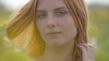Kırmızı saçlı ve yeşil gözlü kendine güvenen şehvetli kızın kameraya bakarak ayçiçeği tarlasında duran yakın çekim portresi. Güzellik kavramı, doğayla bağlantı. Yavaş çekim.