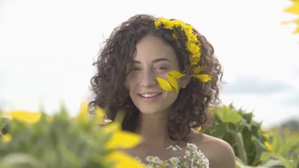 Kır çiçeği buketi ile ayçiçeği alanında ayakta gülümseyen kameraya bakan eğlenceli bir kıvırcık kız portresi. Parlak sarı renk. Özgürlük kavramı. Dışarıda mutlu bir kadın. Yavaş çekim. — Stok video