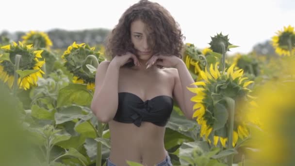 Portrait of a beautiful curly girl in black bra posing and touching her hair standing on the sunflower field. Bright yellow color. Freedom concept. Happy woman outdoors. Slow motion — Stock Video