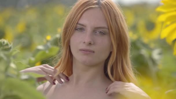 Portrait of young confident girl with red hair and green eyes looking at the camera touching her long hair standing on sunflower field. Concept of beauty, connection with nature. Slow motion — Stock Video