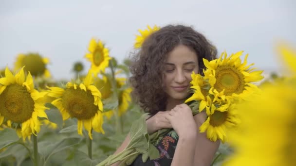 花の花束を持ってヒマワリ畑に立つ美しい巻き毛の女の子の肖像画。明るい黄色。スローモーション — ストック動画