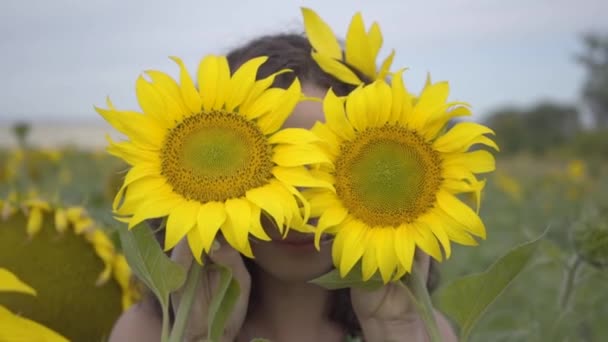 Portret pięknej kręcone dziewczyny patrząc na aparat uśmiechnięty obejmujące jej twarz z dwoma słoneczniki w polu słonecznika. Jasny żółty kolor. Połączenie z naturą. Życie na wsi. Zwolnionym — Wideo stockowe