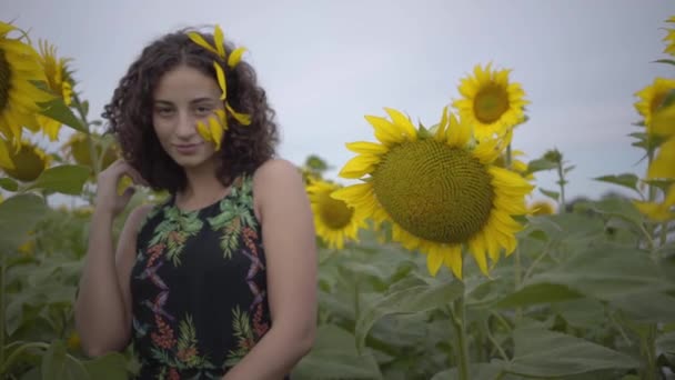 Porträt einer lieblich lustigen lockig verspielten sinnlichen Frau, die lächelnd in die Kamera im Sonnenblumenfeld blickt. leuchtend gelbe Farbe. Freiheitsbegriff. glückliche Frau im Freien. Zeitlupe. — Stockvideo