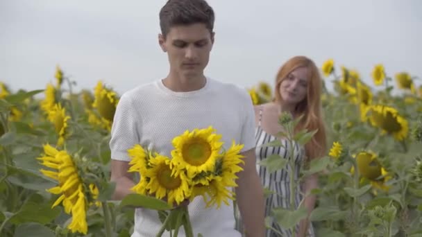Młoda kobieta i mężczyzna bawią się na zewnątrz. Facet trzyma słoneczniki na pierwszym planie, podczas gdy jego dziewczyna stoi w tle. Romantyczna relacja. Życie na wsi — Wideo stockowe