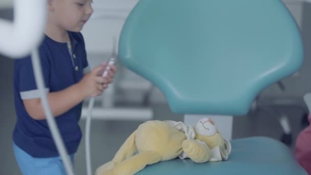 Imádnivaló kisfiú játszik az orvosi eszközök ül a széken a fogorvosi rendelő. Gondtalan gyermek látogató orvos. Fiú játszik a fogorvoshoz. Fogászati kezelés, gyógyászati koncepció. Fogászati ellátás. — Stock videók