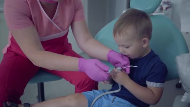 医療用マスクと手袋の歯科医は、歯科医院の椅子に座っている小さな気楽な男の子の歯をチェックする準備ができています。職場の女性専門医の胃科医。歯科治療. — ストック動画