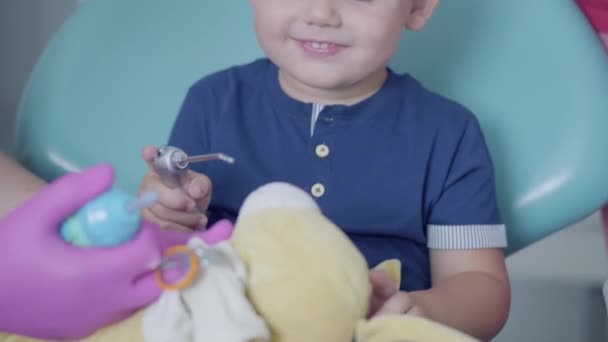 Um rapaz despreocupado sentado na cadeira no consultório dentário. Bonito criança brincando com brinquedo de pelúcia. Tratamento dentário, conceito médico . — Vídeo de Stock