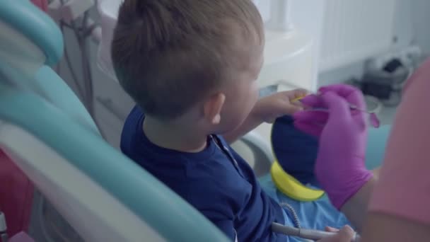 Mani da dentista guantate che controllano i denti del ragazzino spensierato seduto sulla sedia mostrandogli uno specchio da vicino. Stomatologia, trattamento, concetto medico. Cure dentali . — Video Stock