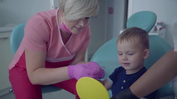 Fogorvos orvosi maszkot és kesztyűt kész ellenőrző fogas kis gondtalan fiú ült tükör a széken. Női szakmai orvos stomatologist a munkahelyen. Fogászati kezelés, orvosi koncepció — Stock videók