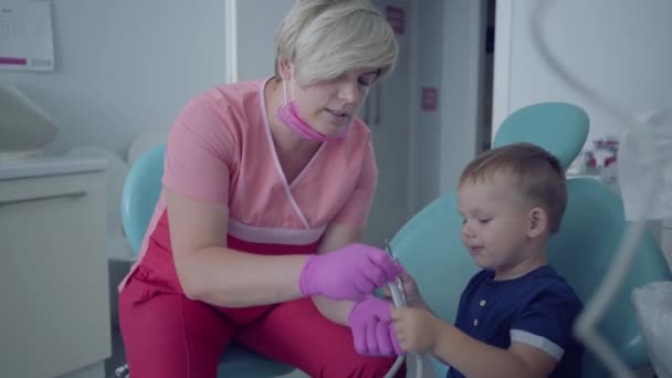 Tıbbi maske ve eldiven diş hekimi sandalyede oturan küçük kaygısız çocuk dişlerini kontrol etmeye hazır. Kadın profesyonel doktor stomatoloji uzmanı iş başında. Diş tedavisi, tıbbi kavram. — Stok video