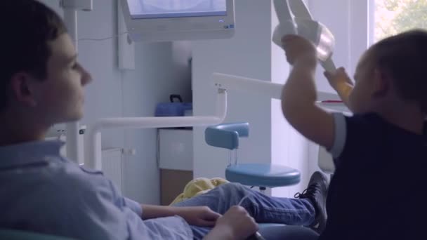 치과 의사 사무실의 의자에 앉아있는 십대 소년은 그의 동생이 의사를 모방 램프를 재생하는 동안. 의사 사무실에서 놀고있는 아이들. 치과학, 의학 개념 — 비디오