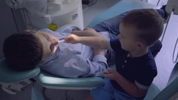 Little boy playing a doctor and teenage guy sitting in the chair in dentist office. Child inspecting teeth of boy. Kid playing a doctor. Dental treatment, medical concept. Dental care. — Stock Video