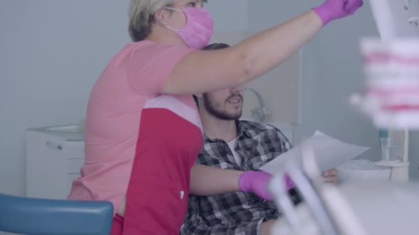 Dentiste féminine en masque rose et gants montrant au patient masculin photo de ses dents à l'écran. Le jeune homme rend visite au médecin. Traitement dentaire, concept médical. Soins dentaires . — Video