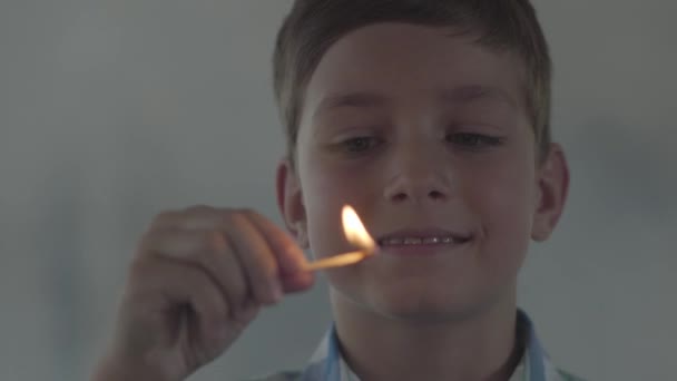 暗い煙の部屋で試合で遊ぶ小さな男の子のクローズアップ顔。その子は笑顔で試合に火をつけた。子供は火遊びをする。放火未成年者 — ストック動画