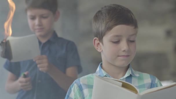 Schattige kleine tweelingbroers in de rokerige kamer. Een jongen verlicht het papier met een lichtere en een ander kind lezen op de voorgrond. Brandstichting. — Stockvideo