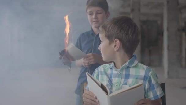 Twee kleine tweelingbroers in de rokerige kamer. Een jongen verlicht het papier met een lichtere en een ander kind lezen op de voorgrond. Brandstichting. — Stockvideo