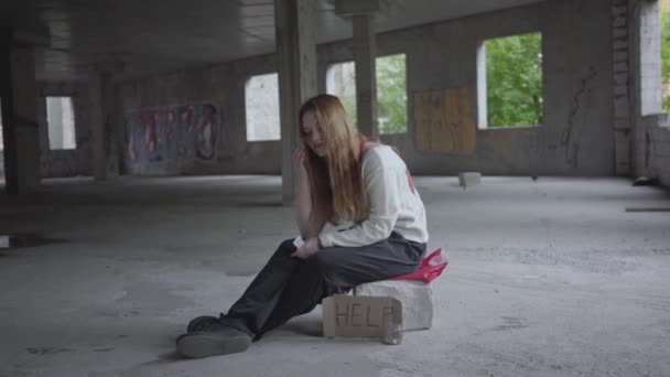 Caucasienne jeune fille triste assis dans un bâtiment abandonné avec un carton aide signe en attente de soutien et d'aide . — Video