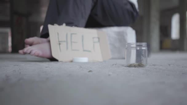 Feet of barefoot poor girl on concrete floor. A blurred sign that says help and jar with coins lying in the foreground. People pass by, one man stops and puts money in the jar. Helping the poor — Stock Video