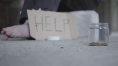 Beton zeminde yalınayak zavallı kız ayakları. Ön planda yatan sikkeler ile yardım ve kavanoz yazan bulanık bir işaret. İnsanlar geçip, bir adam durup kavanoza para koyar. Yoksula yardım etmek