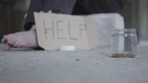 Feet of barefoot poor girl on concrete floor. A blurred sign that says help and jar with coins lying in the foreground. People pass by, one man stops and puts money in the jar. Helping the poor — Stock Video