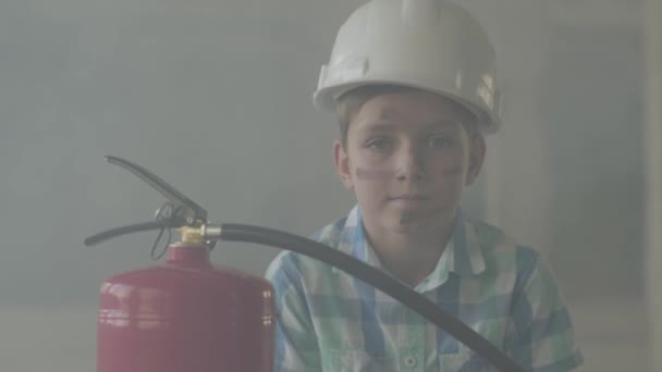 煙の背景にカメラを見て消火器と白い保護ヘルメットの小さな男の子の肖像画. — ストック動画