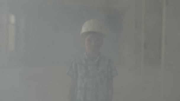 Портрет симпатичного мальчика в защитном шлеме, смотрящего в камеру на фоне дыма в помещении. Концепция пожара, катастрофы, огнестойкости, несоблюдения правил безопасности . — стоковое видео