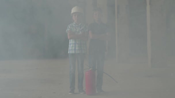 Dos niños pequeños con cascos blancos de seguridad están junto a un extintor de incendios en una habitación humeante. Concepto de incendio, inflamabilidad, incumplimiento de las normas de seguridad . — Vídeo de stock