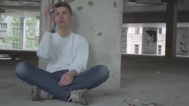 Terk edilmiş binada beton zeminde oturan genç yalnız adam elinde beyaz hap tutuyor, sonra da yutuyor. Kamera eğilir, döner. Bağımlı uyuşturucu alma — Stok video