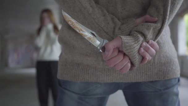 Zblízka, pohled na ozbrojeného muže, který drží nůž v dlaních a skrývá ho za zády. Vystrašená mladá žena stála na pozadí před vrahem. Koncept násilí, vraždy — Stock video