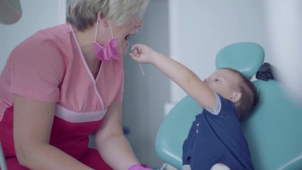 Dentista in maschera medica e guanti pronti a controllare i denti di un bambino spensierato seduto sulla sedia. Il bambino cattivo che gioca con il dottore. Trattamento dentale, concetto medico . — Video Stock