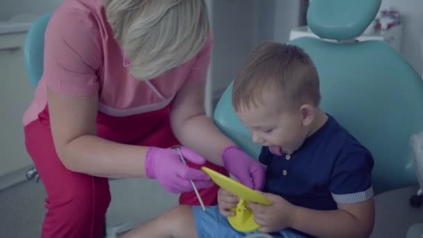 Стоматолог в медицинской маске и перчатках, готовый проверить зубки маленького беззаботного мальчика, сидящего с зеркалом в кресле. Женщина профессиональный врач стоматолог на работе. Лечение зубов, медицинская концепция — стоковое видео
