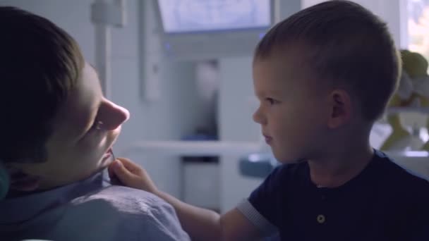Nastoletni chłopiec siedzi na krześle w gabinecie stomatologicznym, podczas gdy jego mały brat gra z lekarzem, kontrolując zęby z bliska lustro. Leczenie stomatologiczne, koncepcja medyczna. Opieka stomatologiczna. — Wideo stockowe