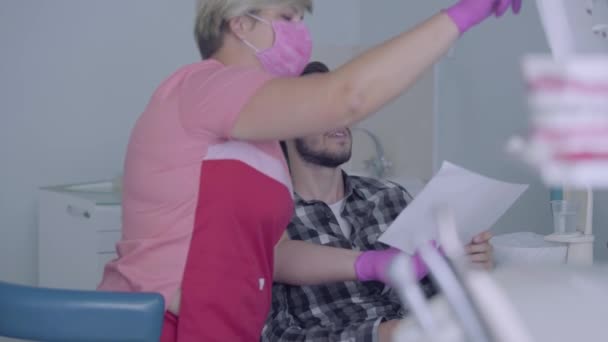 Pembe maskeli kadın diş hekimi ve erkek hastaya dişlerini gösteren eldivenler ekranda. Doktoru ziyaret eden genç adam. Diş tedavisi, tıbbi konsept. Diş bakımı. — Stok video